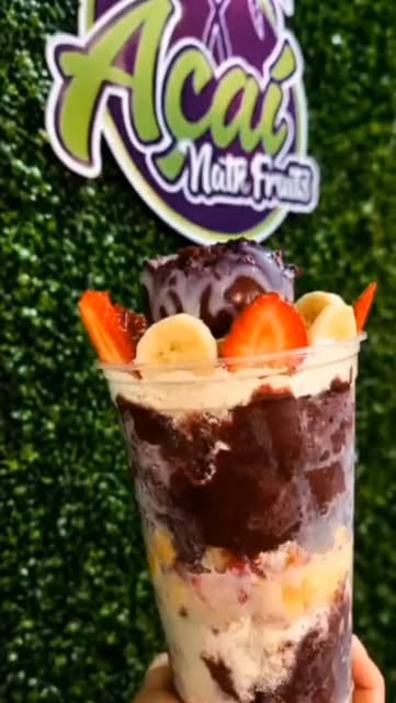 Copo de açaí em camadas com creme, morango e banana, na frente do painel verde com o logo